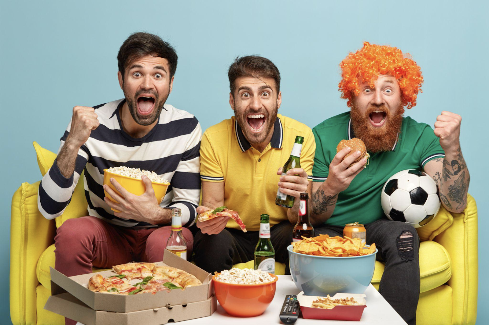 Três homens comendo pizza e bebendo cerveja enquanto assistem uma partida esportiva em cenário colorido