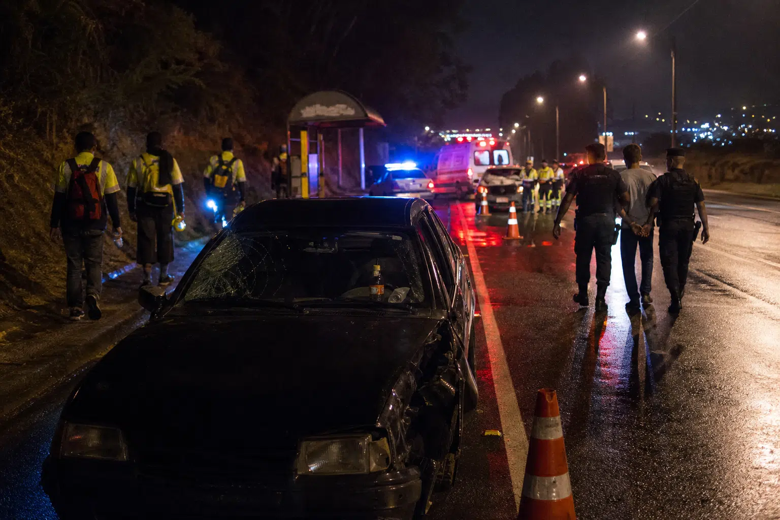 Carro danificado em acidente numa estrada molhada à noite, com socorristas, ambulância e cones de sinalização