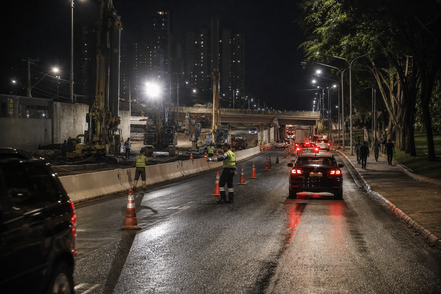 Obras noturnas em avenida molhada, com máquinas, trabalhadores, cones, carros e trânsito sob um viaduto